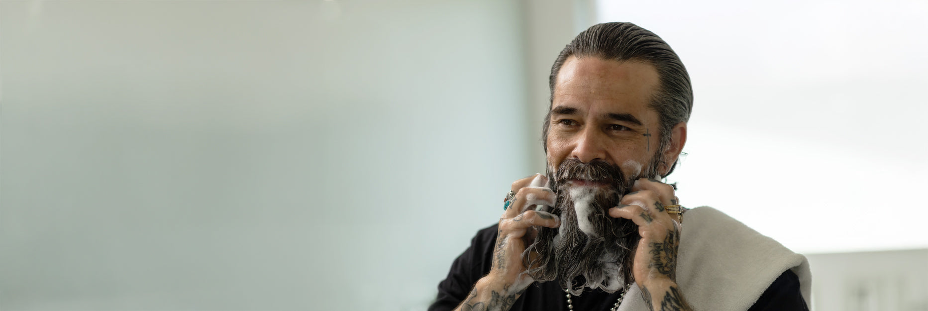 A man with a dark beard and slicked-back hair lathering soap onto his beard, wearing a black shirt and a white towel draped over his shoulder, set against a bright, softly lit background.